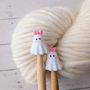 May include: Two white ghost-shaped stitch markers with pink bows sit atop wooden knitting needles, set against a backdrop of a large, cream-colored yarn ball. The markers have black dot eyes and pink blush. The needles are positioned in front of the yarn.