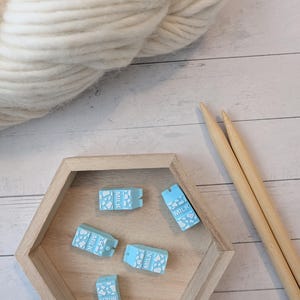 May include: A light blue milk carton-shaped stitch markers with white paw prints and the word "MILK" are in a wooden hexagon tray. A large ball of white yarn and two wooden knitting needles are also in the image.
