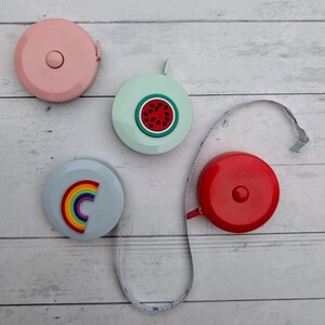Puede incluir: Cuatro cintas m&eacute;tricas retr&aacute;ctiles de colores con diferentes dise&ntilde;os. Las cintas m&eacute;tricas son rosa, azul claro, verde claro y rojo. Los dise&ntilde;os son una sand&iacute;a, un arco&iacute;ris, un color s&oacute;lido y un color s&oacute;lido.