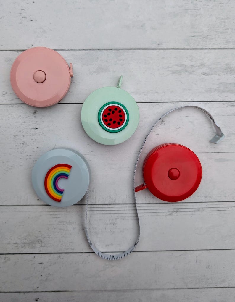 Puede incluir: Cuatro cintas m&eacute;tricas retr&aacute;ctiles de colores con diferentes dise&ntilde;os. Las cintas m&eacute;tricas son rosa, azul claro, verde claro y rojo. Los dise&ntilde;os son una sand&iacute;a, un arco&iacute;ris, un color s&oacute;lido y un color s&oacute;lido.