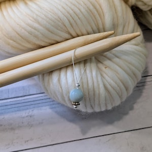 May include: A light blue bead with gold accents is threaded onto a silver needle, which is resting on top of a ball of white yarn and two wooden knitting needles.