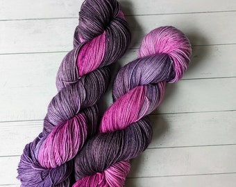 May include: Two skeins of hand-dyed yarn in shades of purple, pink, and lavender. The yarn is arranged in a loose, twisted shape.