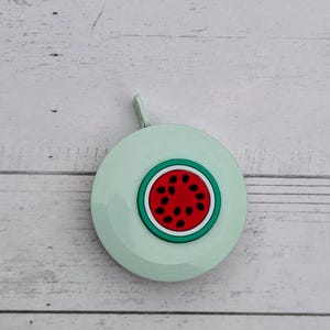 Puede incluir: Una cinta m&eacute;trica retr&aacute;ctil de color verde claro con un dise&ntilde;o de sand&iacute;a. La sand&iacute;a es roja con semillas negras y una c&aacute;scara verde.