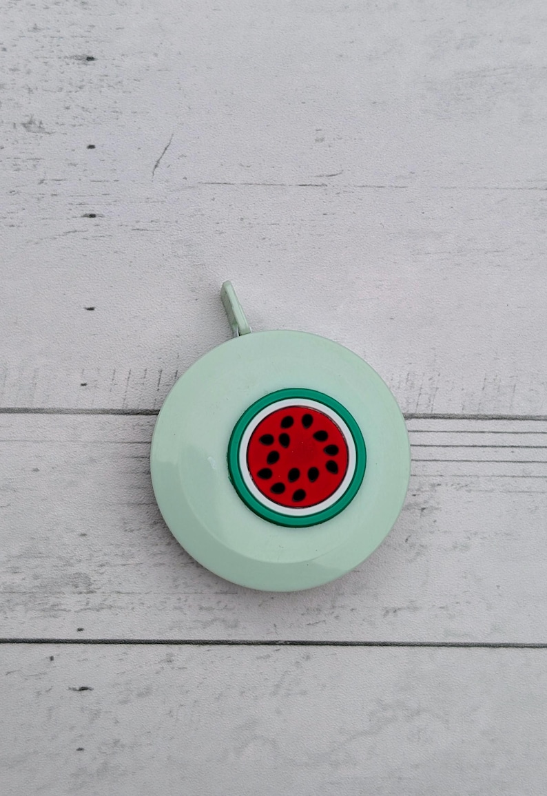 Puede incluir: Una cinta m&eacute;trica retr&aacute;ctil de color verde claro con un dise&ntilde;o de sand&iacute;a. La sand&iacute;a es roja con semillas negras y una c&aacute;scara verde.