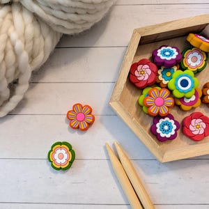 May include: A collection of small, multi-colored flower-shaped beads in a hexagonal wooden tray. The beads feature vibrant petals in shades of red, purple, green, orange, and blue. Two wooden knitting needles and a ball of white yarn are present.