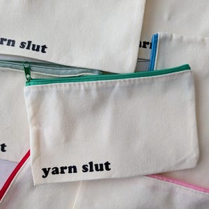 May include: Off-white canvas zipper pouches with various colored zippers and trim. The text "yarn slut" is printed in black on the front of one pouch. Other pouches are visible with similar text. The pouches are designed for storing craft supplies.