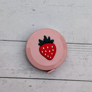 Puede incluir: Una cinta m&eacute;trica retr&aacute;ctil rosa con un dise&ntilde;o de fresa. La fresa es roja con semillas blancas y hojas verdes.