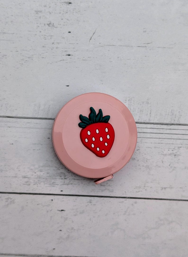 Puede incluir: Una cinta m&eacute;trica retr&aacute;ctil rosa con un dise&ntilde;o de fresa. La fresa es roja con semillas blancas y hojas verdes.