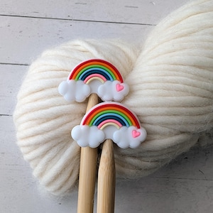 May include: Two rainbow-themed stitch markers with white clouds and pink hearts rest on wooden knitting needles, set against a backdrop of a large ball of white yarn. The rainbows feature vibrant colors.