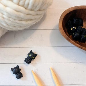 May include: Small black cat-shaped buttons with pointed ears and yellow eyes. One button is shown from the front with visible facial features, while another is shown from the back. These buttons are matte black and have a smooth, molded surface.
