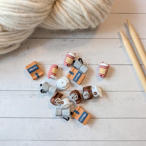 May include: A collection of coffee-themed charms, including coffee cups, coffee bags, and coffee makers, scattered on a white wooden surface. A ball of cream-colored yarn and two wooden knitting needles are also present. The charms have the word "Coffee" on them.