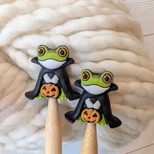May include: Two bamboo knitting needles topped with decorative frog figures. Each green frog wears a black cape and holds a small orange jack-o'-lantern. The frogs have large orange eyes and white chest details, attached to the light brown wooden shafts.