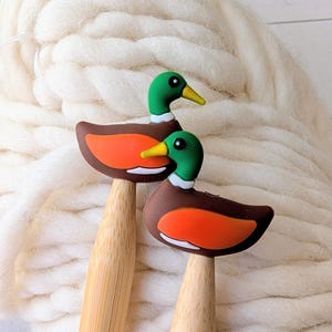 May include: Two bamboo knitting needles featuring mallard duck toppers. Each duck has a green head, a yellow beak, a white neck ring, and a brown body with a bright orange wing patch. The needles are smooth, light-colored wood with a tapered tip.