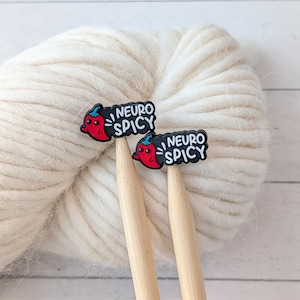 May include: Two knitting needles with "NEURO SPICY" charms featuring a red chili pepper design. The needles are inserted into a ball of thick, off-white yarn. The charms have a black background and white text.