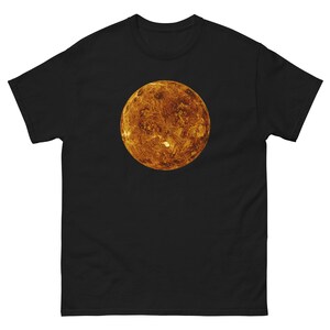 May include: A black short-sleeve t-shirt featuring a centered, circular graphic of the planet Venus. The planet is depicted in shades of orange, yellow, and brown, showcasing a textured, cratered surface against the solid black fabric background.