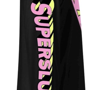 May include: A black long-sleeve shirt featuring the word SUPERSLUT printed vertically down the sleeve in a bold, three-dimensional font with pink lettering and yellow highlights. A partial pink and yellow emblem is visible on the chest area.