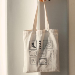 Peut inclure: Sac fourre-tout crème avec des illustrations noires d'un dragon, de coeurs, de livres, d'un nœud, d'une tasse à café, d'une épée et le texte "Everything is Letters". Le sac est tenu par une personne.