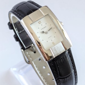 Könnte beinhalten: Eine rechteckige silberne und weiße Uhr mit einem schwarzen Lederarmband. Das Zifferblatt der Uhr hat einen weißen Hintergrund mit schwarzen Markierungen und das Wort "YEMA" ist in Schwarz gedruckt. Das Zifferblatt der Uhr zeigt auch "QUARTZ" in Schwarz an.
