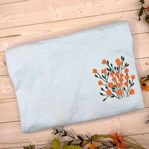 May include: A light blue folded t-shirt with an embroidered floral design. The embroidery features orange flowers and green stems. The shirt is made of a soft material and is folded neatly.