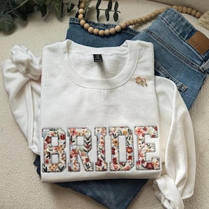 May include: White sweatshirt with the word "BRIDE" spelled out in floral fabric. The letters are outlined in white and have a vintage floral pattern.