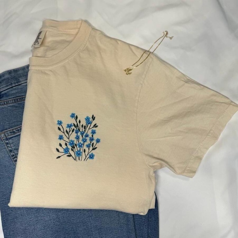 May include: A cream-colored t-shirt with a blue floral embroidery design. The shirt is paired with blue denim jeans and a gold necklace. The t-shirt features a crew neck and short sleeves. The floral design is centered on the chest.