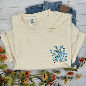 May include: Cream-colored Comfort Colors t-shirt with a small embroidered bouquet of blue flowers. A gold necklace with a pendant rests on the shirt, which is paired with blue jeans. The shirt is size L-G.