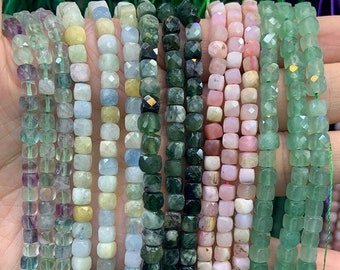 May include: Assortment of faceted gemstone beads in various colors, including green, blue, pink, and yellow. The beads are square shaped and strung on a thread.