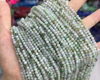 May include: Strands of small, round, faceted beads in shades of pale green and white. The beads are held in a hand, showcasing their delicate texture and subtle color variations. The beads are likely for jewelry making.