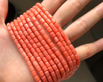May include: Several strands of coral-colored beads, each composed of small, rectangular segments. The beads are held in a hand, showcasing their color and texture. The beads appear to be strung together, forming multiple bracelets.