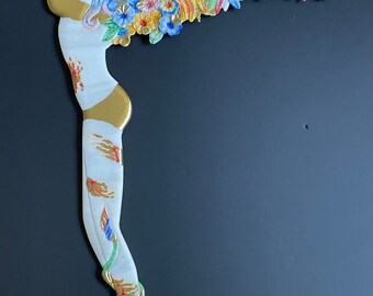 May include: A white ceramic sculpture of a woman with wings made of flowers and gold accents. The woman is standing with her arms raised above her head and her wings are spread out behind her. The sculpture is painted in shades of blue, pink, yellow, and green.