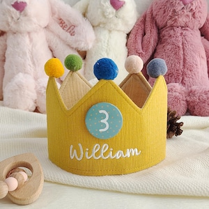 May include: Yellow felt crown with colorful pom poms, a blue and white circle with the number 3 and the name William embroidered on it. The crown is sitting on a white surface with stuffed animals in the background.