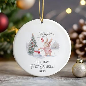 May include: A white ceramic Christmas bauble featuring a watercolour illustration of a bunny in a pink sweater building a snowman. The bauble has the text "Sophia's First Christmas 2025" in black script and a gold hanging cord.