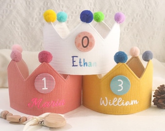 May include: Three felt crowns with pom-poms on top. The crowns are white, pink, and yellow. Each crown has a number and a name embroidered on it. The numbers are 0, 1, and 3. The names are Ethan, Maria, and William.