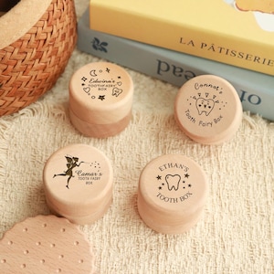 May include: Four small, round wooden tooth fairy boxes. Each box is personalised with a name and features a tooth and star design. The boxes are light brown and sit on a cream-coloured surface.