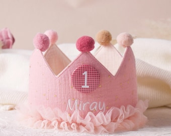Op de afbeelding: Een roze stoffen kroon met een gefranjerde rand, versierd met pompons en gouden spikkels. Een cirkelvormige patch toont het nummer "1" en de naam "Miray" is eronder geborduurd. Een feestelijk accessoire voor een eerste verjaardag.
