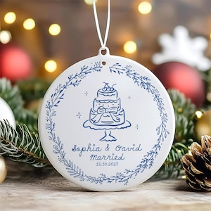 Könnte beinhalten: Weißer Hochzeitsornament aus Keramik mit einer blauen Illustration einer mehrstöckigen Torte, umgeben von einem floralen Kranz. Der Ornament ist mit den Namen "Sophia & David" und dem Datum "12.10.2025" personalisiert. Eine weiße Schnur ist zum Aufhängen befestigt.