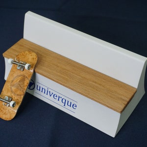 以下が含まれることがあります： 白と木目調のベンチにミニチュアスケートボードが置かれています。スケートボードは明るい茶色の木目模様です。ベンチには青色で「Univerque」の文字が印刷されています。