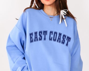 May include: A Carolina blue sweatshirt with "EAST COAST" printed across the chest. The model is wearing red and white checked shorts and black boots. The text "Carolina Blue" is in a blue circle.