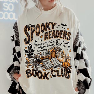 May include: An off-white t-shirt featuring a graphic print with the text "SPOOKY READERS BOOK CLUB" and "Running on coffee, horror & chapters". The design includes illustrations of stacked books, a potted plant, candles, and a jack-o'-lantern.