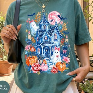 K&ouml;nnte beinhalten: Ein blaugr&uuml;nes Kurzarm-T-Shirt mit einem Grafikdruck eines blauen Spukhauses, zwei Geistern mit rosa Schleifen, orangefarbenen K&uuml;rbissen, bunten Blumen, Flederm&auml;usen und einem Vollmond. Das Shirt wird mit einem wei&szlig;en Faltenrock kombiniert.