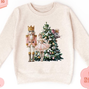 Puede incluir: Sudadera beige con un diseño de Cascanueces, bailarina, árbol de Navidad y ratón. La parte superior de manga larga y cuello redondo tiene el texto "13 COLOR OPTIONS" y "NATURAL".