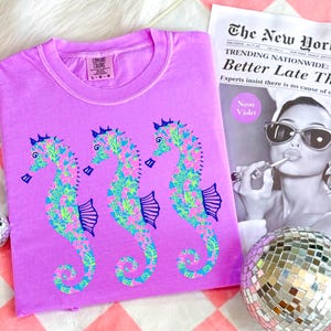May include: A lilac Comfort Colors t-shirt featuring three seahorse designs. The seahorses are decorated with a floral pattern in shades of pink, green, and blue. A newspaper with the headline "Better Late Than" and a disco ball are also in the image.