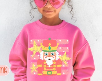 May include: A bright pink long-sleeve sweatshirt featuring a graphic print of a nutcracker wearing a crown, surrounded by yellow stars and white polka dots on a striped background. The sweatshirt is paired with blue denim jeans and heart-shaped glasses.