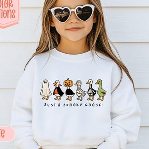 May include: White sweatshirt with a Halloween-themed design. It features cartoon geese in costumes and the phrase "JUST A SPOOKY GOOSE". The image also includes text that says "13 COLOR OPTIONS" and "WHITE".