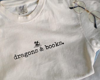 May include: A white t-shirt with a black dragon and the text "dragons & books." The shirt is laying on a white surface with a pair of glasses and blue jeans.