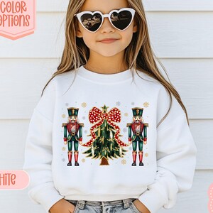 Puede incluir: Sudadera blanca con un diseño navideño que incluye un árbol, un lazo rojo y dos soldados cascanueces. El diseño incluye copos de nieve. El texto "13 COLOR OPTIONS" y "WHITE" son visibles.