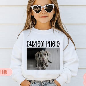 May include: A white sweatshirt with the text "CUSTOM PHOTO" above a grayscale image of a dog. The word "WHITE" is printed in a pink rectangle on the bottom left. The sweatshirt is worn by a person wearing heart-shaped sunglasses.