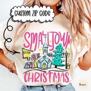 Może przedstawiać: Kremowa koszulka z kolorową grafiką przedstawiającą scenę z małego miasteczka, w tym kościół, domy i choinkę. Tekst "SMALL TOWN CHRISTMAS" jest różowy, z niestandardowym kodem pocztowym. Koszulka jest połączona z jasnoniebieskimi dżinsami.