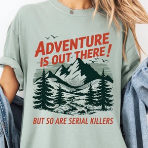 May include: A light green t-shirt with the text "ADVENTURE IS OUT THERE! BUT SO ARE SERIAL KILLERS" in red and black. The graphic features a mountain range, trees, and birds.