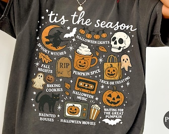 Może przedstawiać: Ciemnoszara koszulka z motywem Halloween. Projekt zawiera wiedźmę na księżycu, czaszkę, dynie, duchy i napis "'tis the season". Inne elementy to nagrobek, kubek do kawy i kaseta.